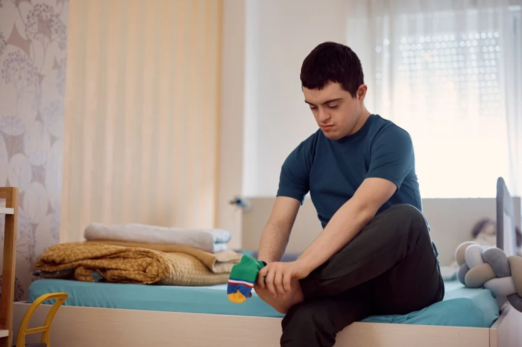 Disabled boy sitting on edge of bed putting socks on